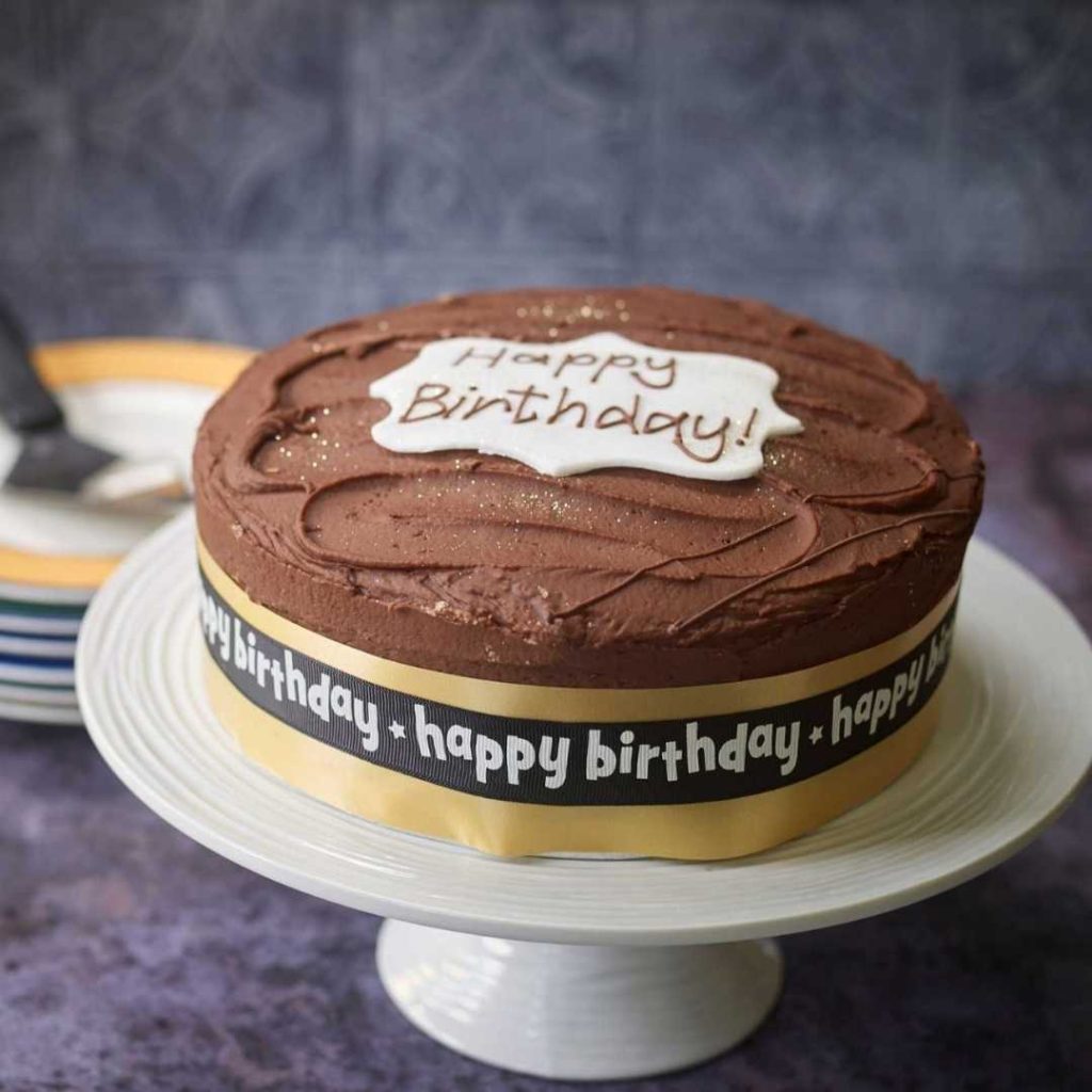 Vegan chocolate birthday cake. Happy birthday piped and happy birthday ribbon. Dairy and egg free birthday cake displayed on a cake stand