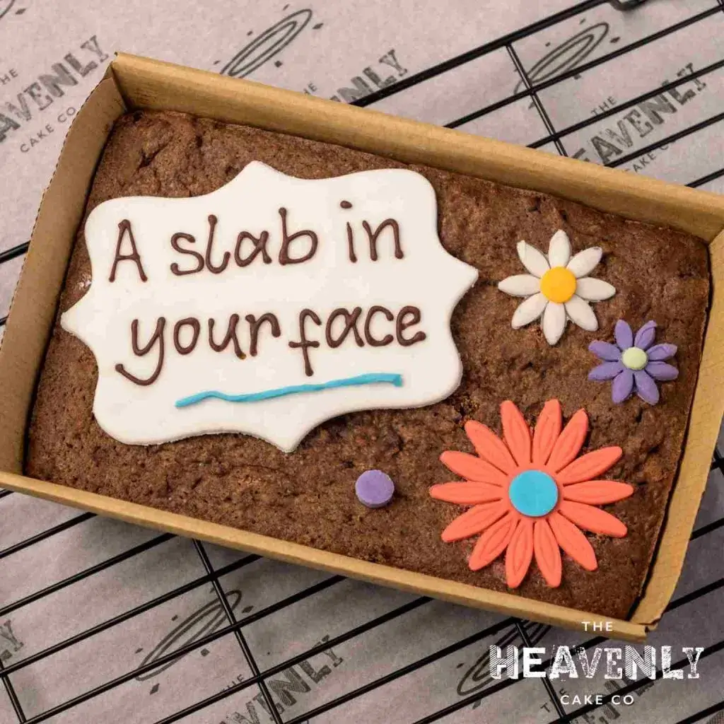 A letterbox brownie in a box with the message "a slab in your face" piped on a white fondant icing disc. Also showing some colourful fondant icing flowers.