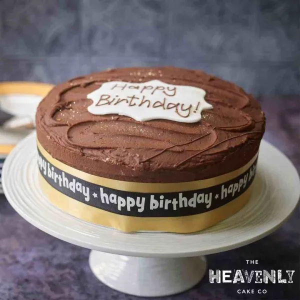 Chocolate birhtday cake displayed on a white cake stand. Happy Birthday piped onto a white disc of fondant icing.